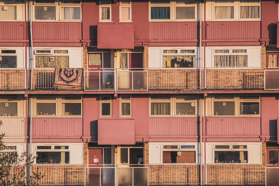 Photograph of an apartment building facade with multiple balconies and windows. The building features pink and cream-colored exterior walls, with some sections having brickwork detail. Several balconi